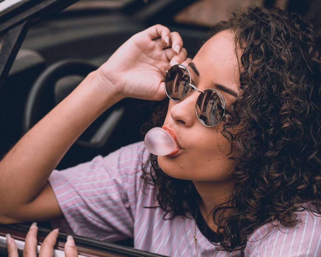 Woman with curly hair and sunglasses blowing gum in car, exuding style and confidence.